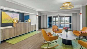 Un área de recepción moderna con un mostrador de madera, un cuadro abstracto de gran tamaño y un espacio de espera con sillas naranjas y una mesa redonda.