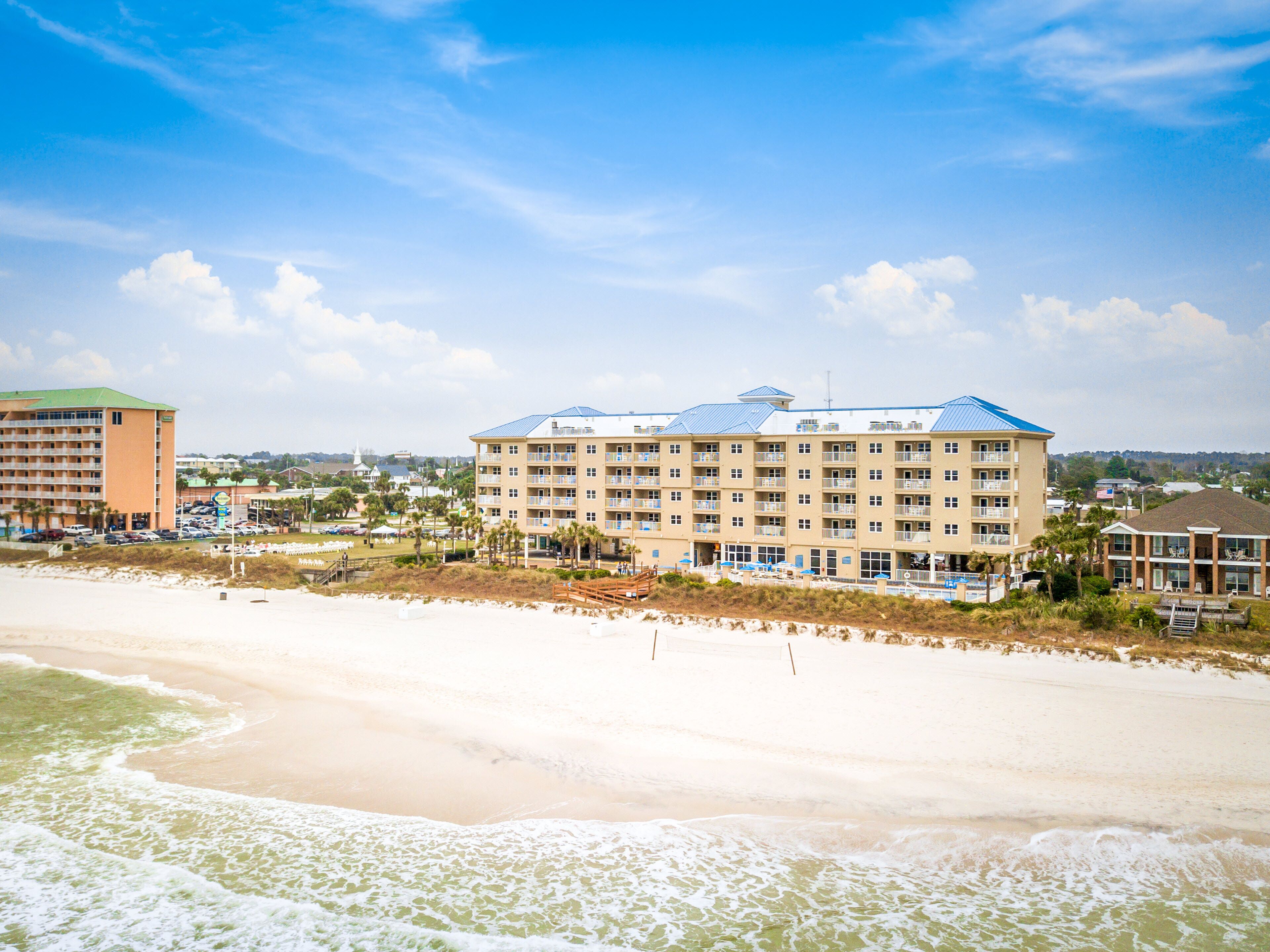 Un hotel frente al mar con varios pisos, una zona de piscina y una playa en primer plano.