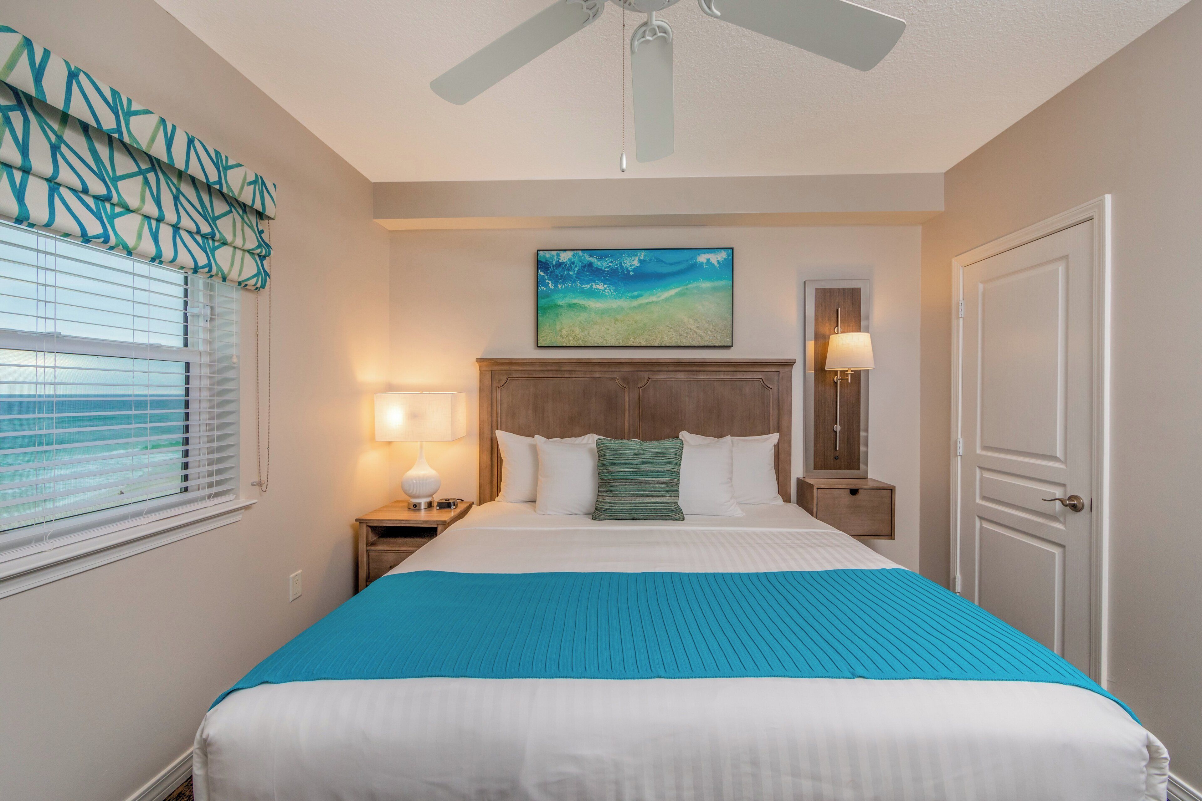 A hotel room with a bed, a ceiling fan, a painting, a nightstand with a lamp, and a view of the ocean through the window.