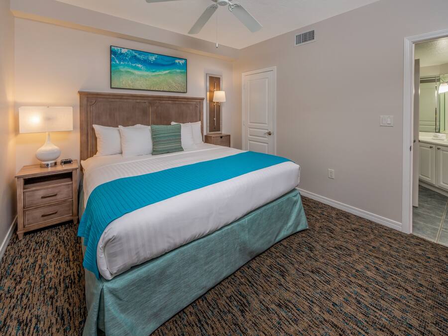 Habitación de hotel con una cama grande, una mesita de noche con lámpara, un cuadro en la pared y un baño visible a través de una puerta abierta.