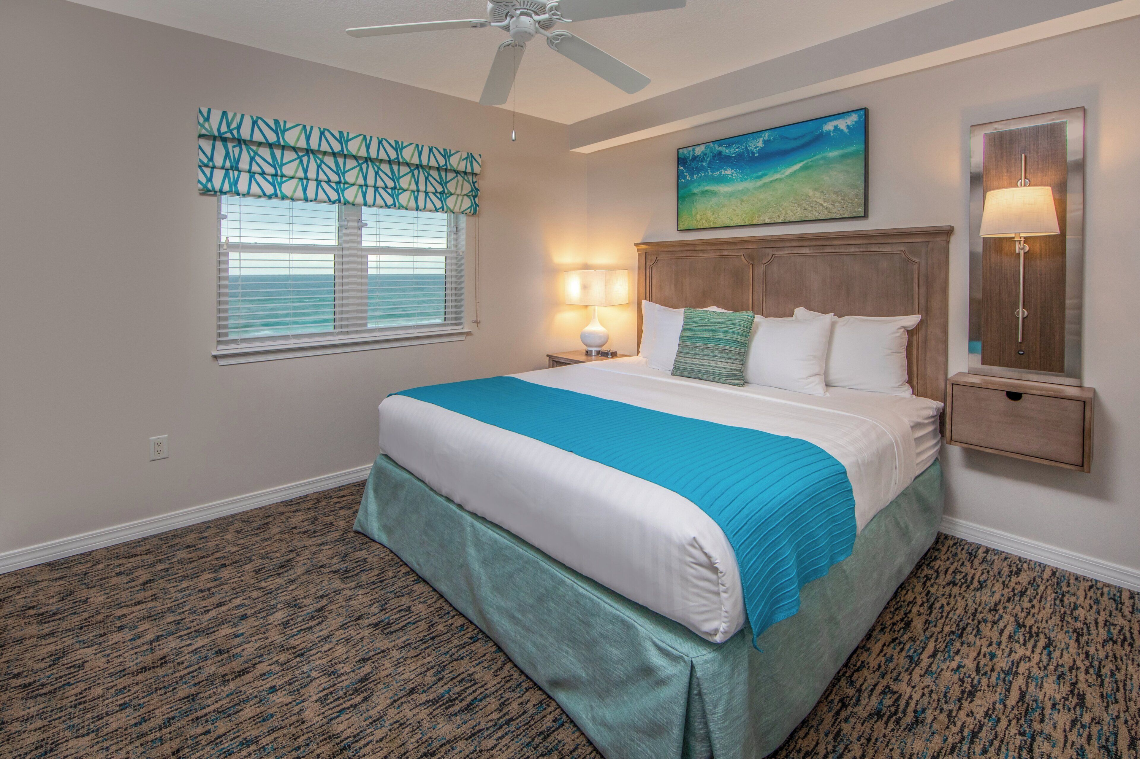 A hotel room with a bed, a nightstand, a lamp, a ceiling fan, a window with blinds, and a painting on the wall.