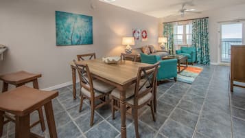 A dining area with a wooden table and chairs, a living room with a teal sofa and a ceiling fan, and a view of the ocean through large windows.
