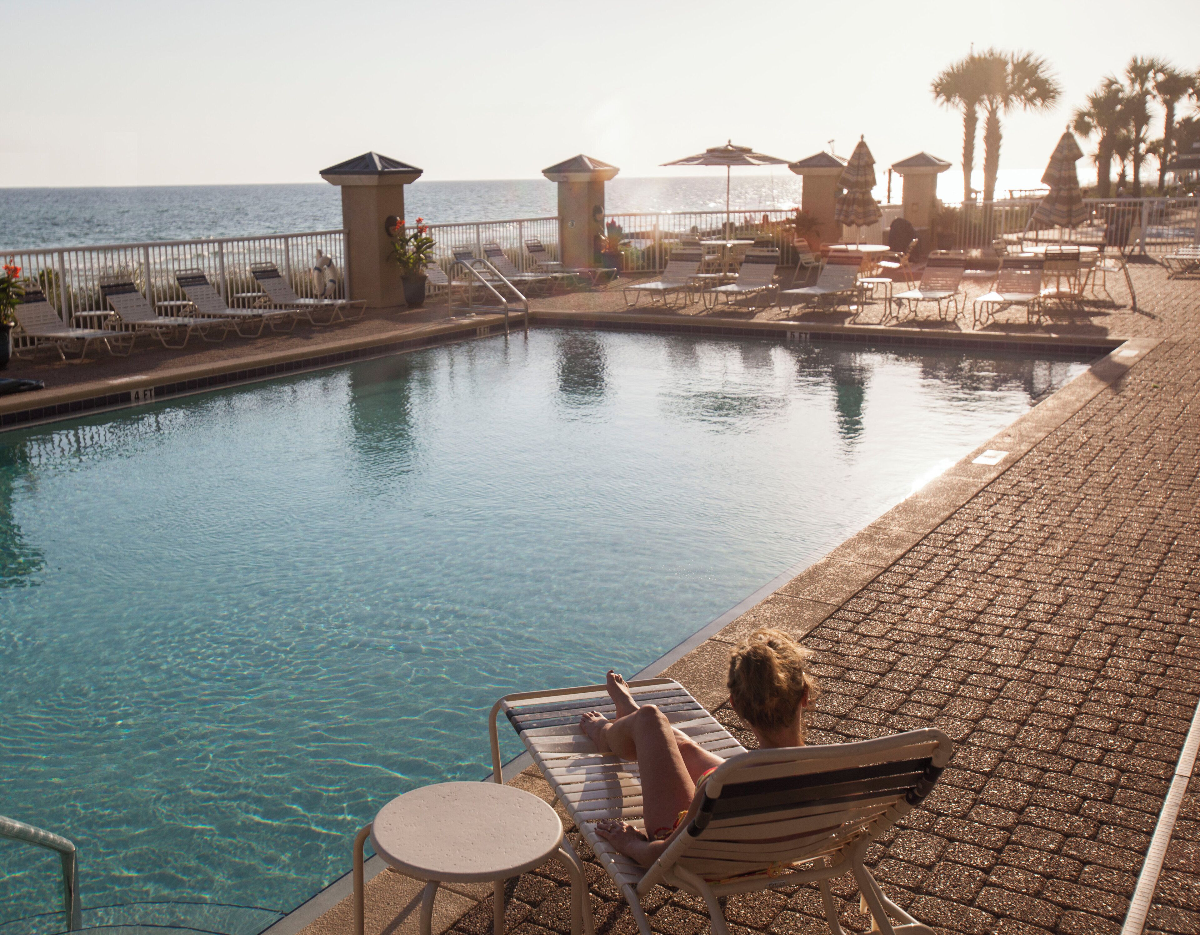A pool area with lounge chairs and umbrellas, a clear pool, and a view of the sea.