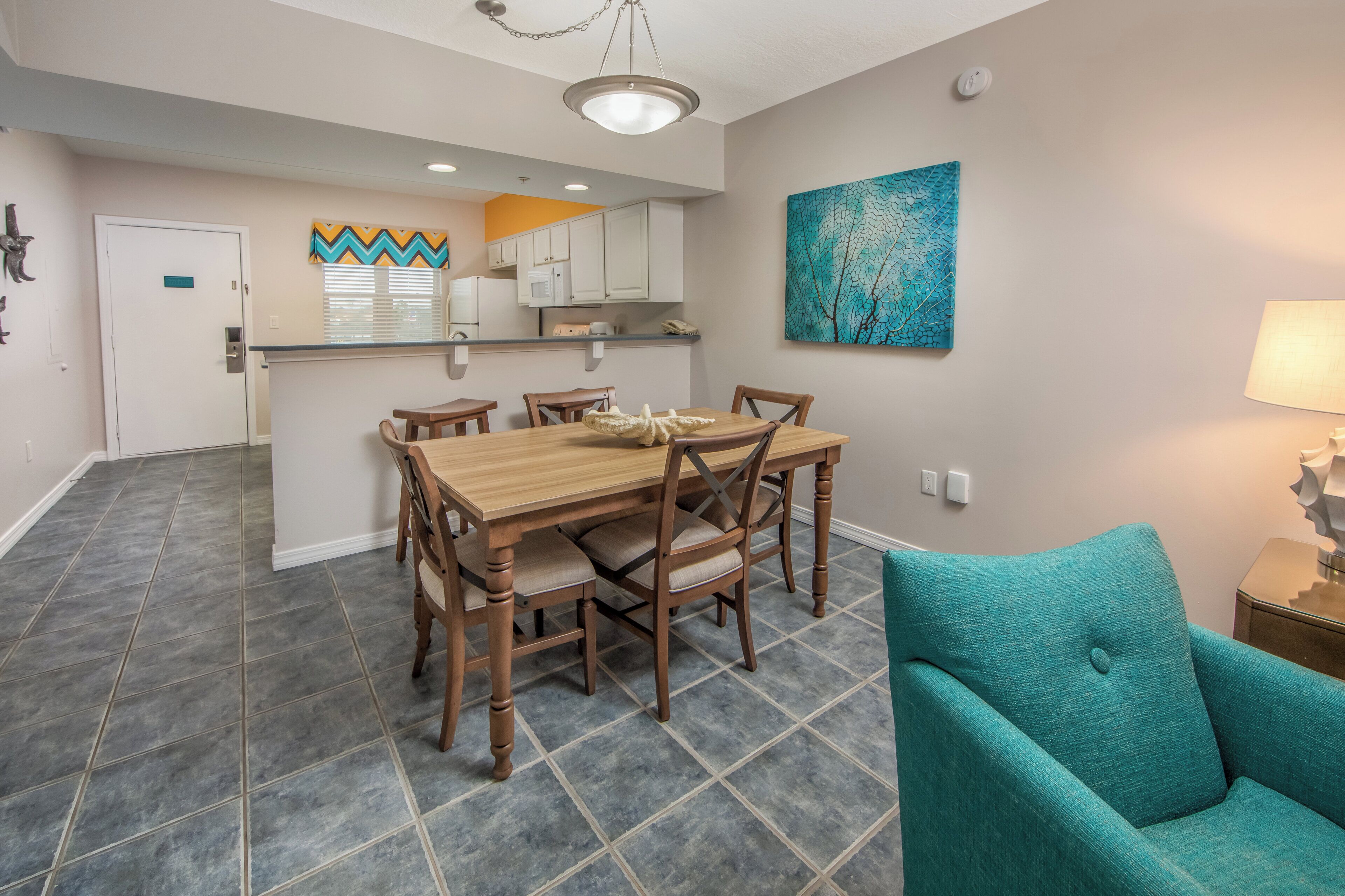 A dining area with a wooden table and chairs, a teal armchair, and a kitchen in the background.