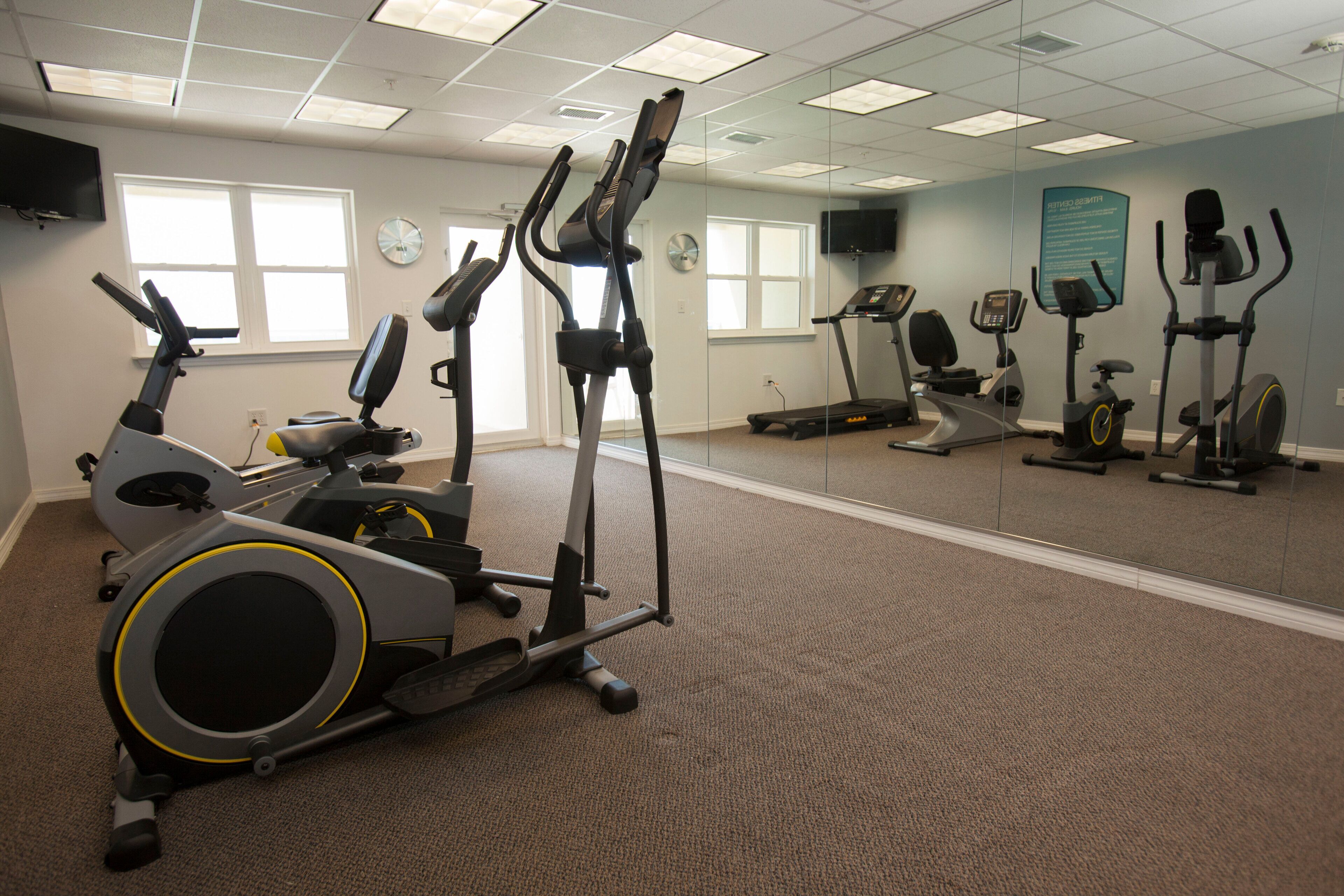 A fitness center with various exercise machines, including treadmills and stationary bikes, arranged in a spacious room with carpeted flooring and large windows.