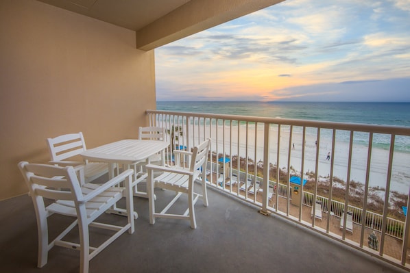Varanda com mesa e cadeiras, vista para uma praia com pessoas e guarda-sóis.