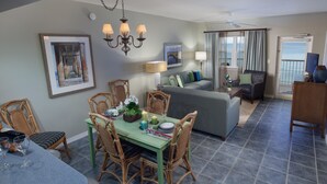 A dining area with a green table and wicker chairs, a grey sofa, and a balcony with curtains.