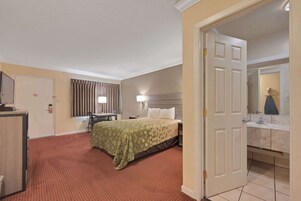 Habitación de hotel con cama, escritorio, silla, baño con lavamanos y espejo, y una puerta que da a otra habitación.