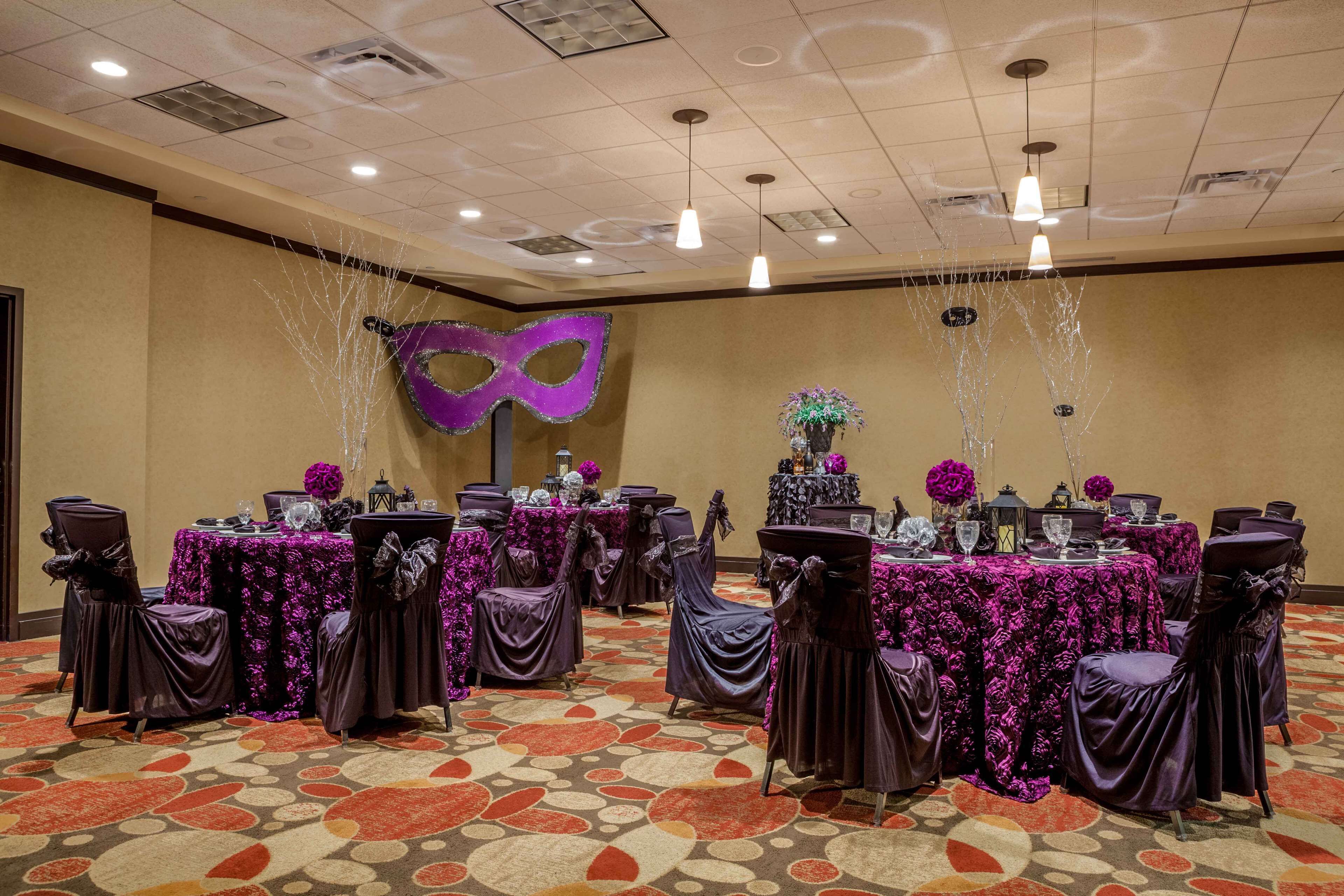 A banquet hall with round tables covered in purple tablecloths, chairs with purple sashes, and a purple mask decoration on the wall.