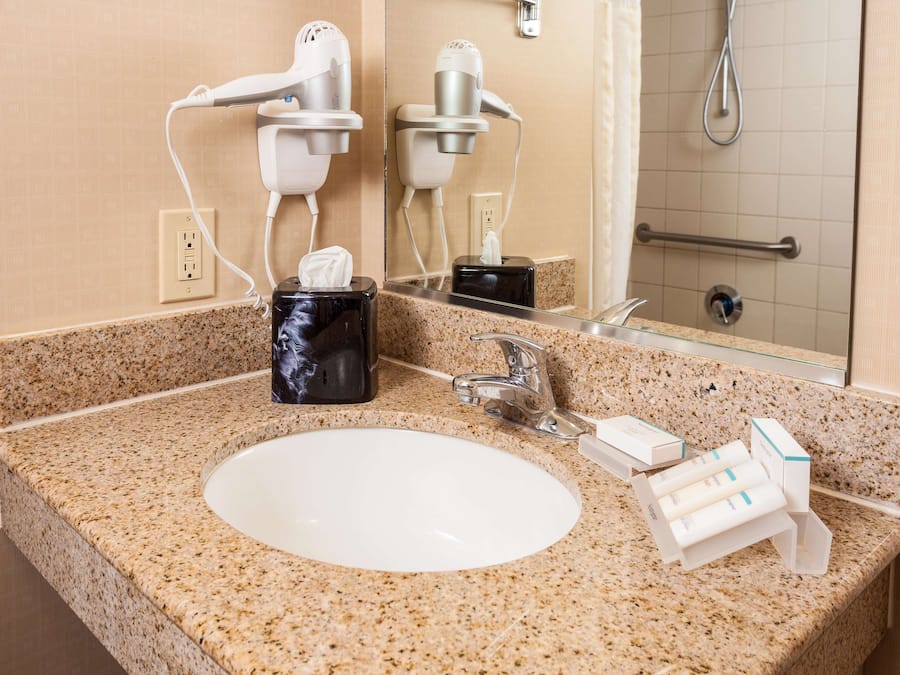 A bathroom with a granite countertop, a white sink, a mirror, a towel dispenser, and two hair dryers mounted on the wall.