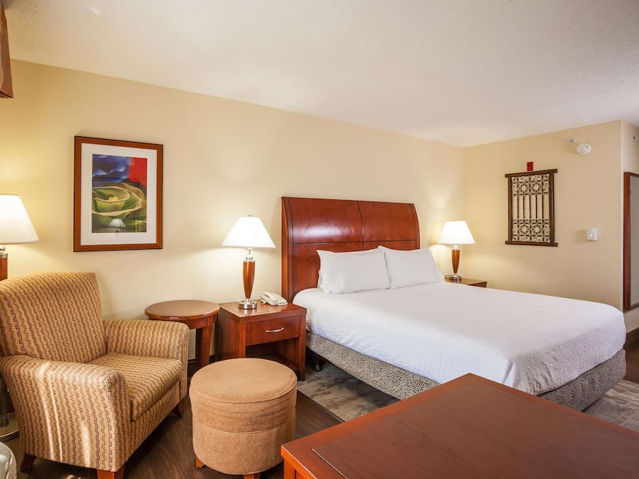 Une chambre d’hôtel avec un lit, une chaise, une table, une lampe, une table de chevet et un tableau au mur.