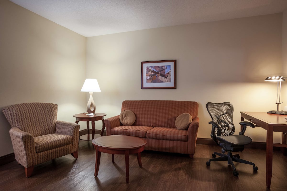 A living room with a sofa, armchair, coffee table, and office chair. There is a lamp on a side table and a framed picture on the wall.