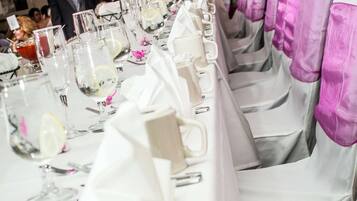 Uma mesa comprida posta para um evento formal, com toalhas brancas e fitas cor-de-rosa nas cadeiras.