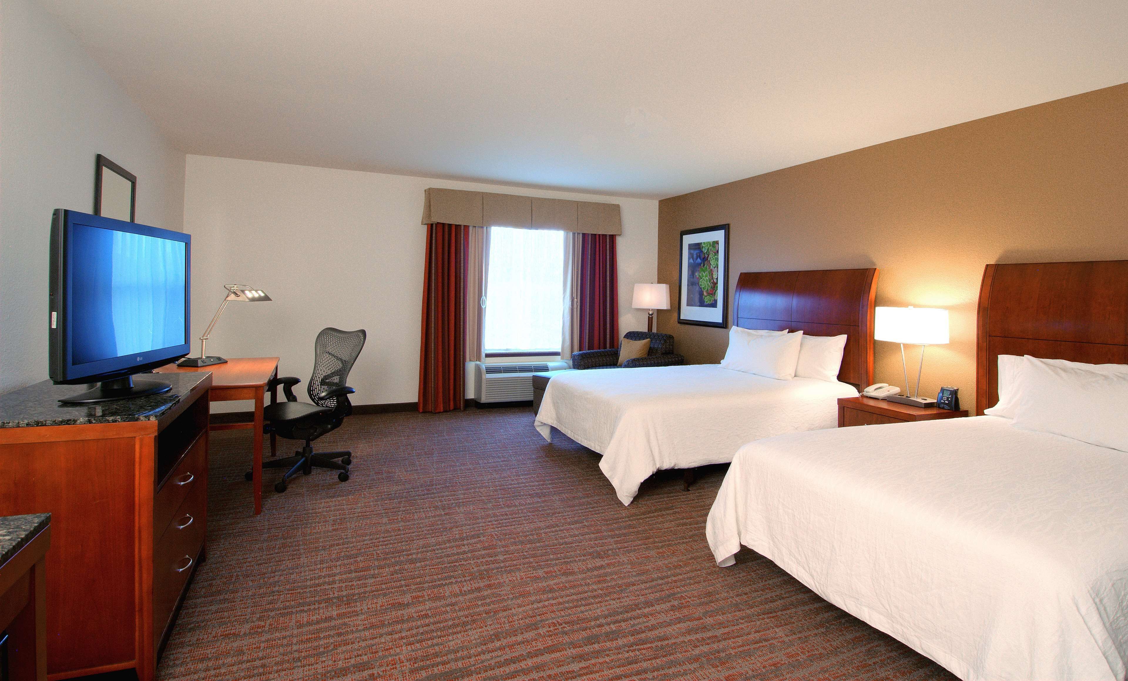 A hotel room with two beds, a desk with a computer, a chair, a TV, and a window with curtains.