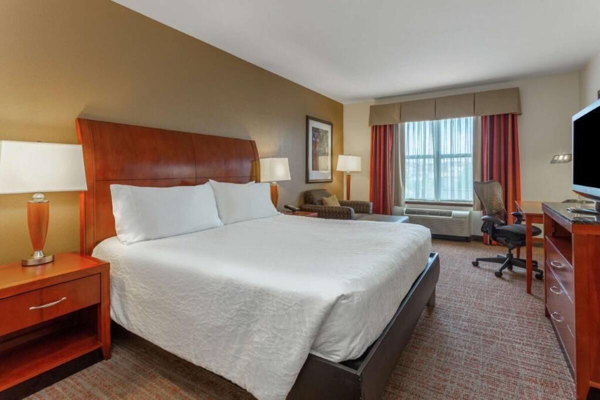 A hotel room with a large bed, a desk, a chair, a television, and a window with curtains.