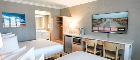 A hotel room with two beds, a desk with a lamp, a television, and a view of a beach.