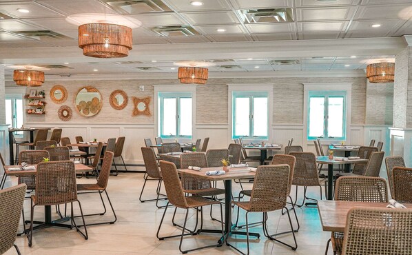 Een ruime eetruimte met rieten stoelen en tafels, grote ramen en decoratieve spiegelwanden.