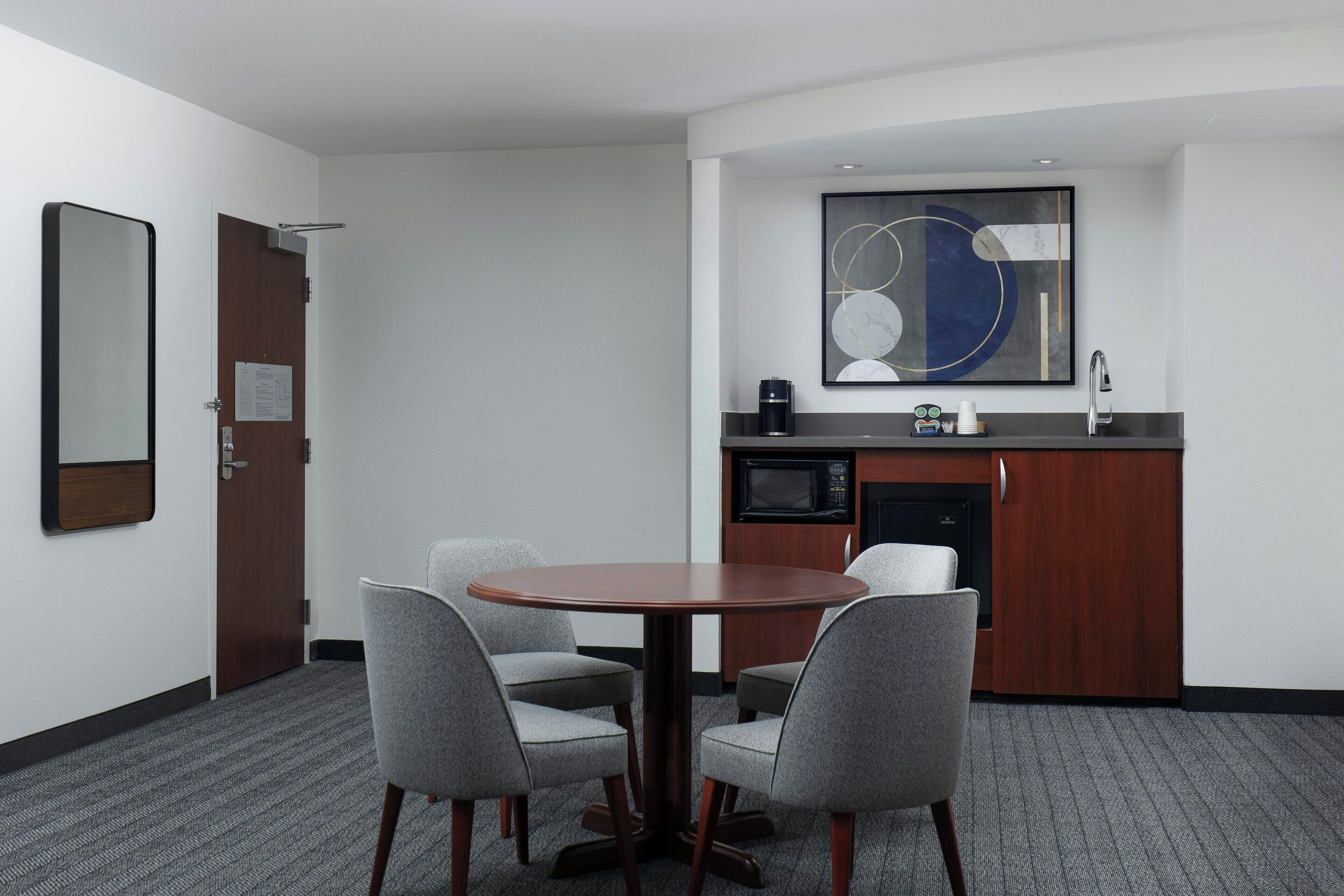A hotel room with a round table, four chairs, a microwave, a coffee maker, a sink, and a large abstract painting on the wall.