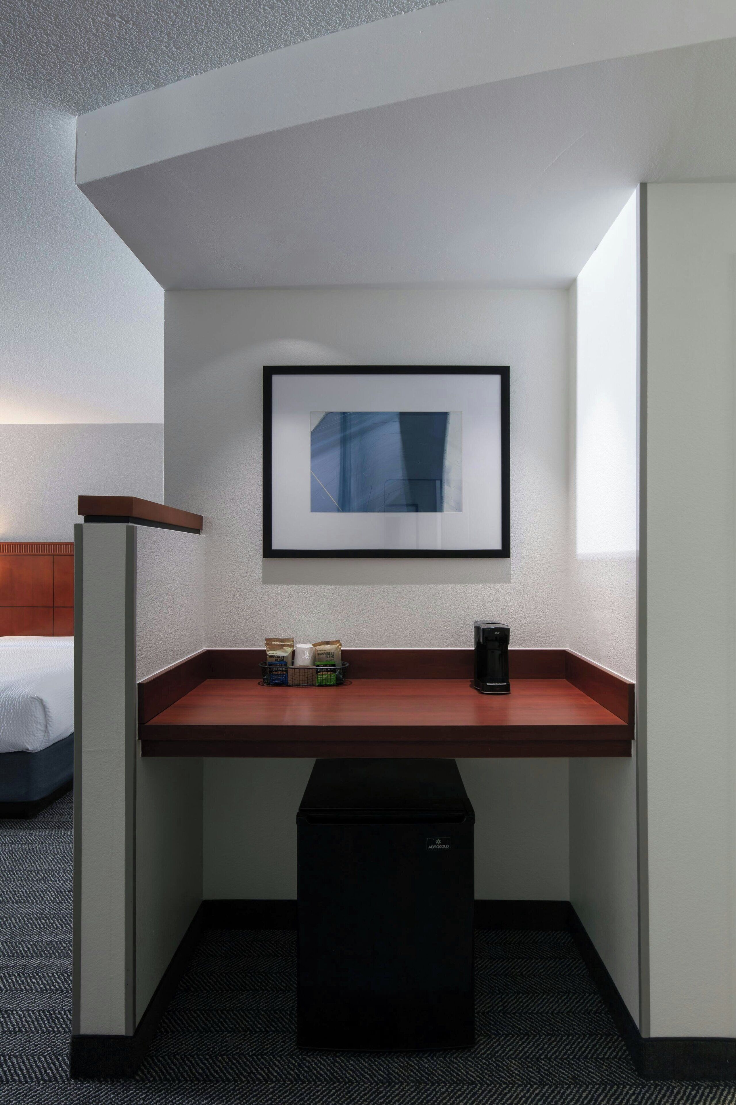 A compact hotel room with a single bed, a small desk with a coffee maker, a framed picture on the wall, and a trash bin.