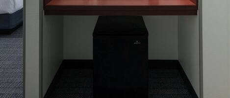 A compact hotel room with a single bed, a small desk with a coffee maker, a framed picture on the wall, and a trash bin.