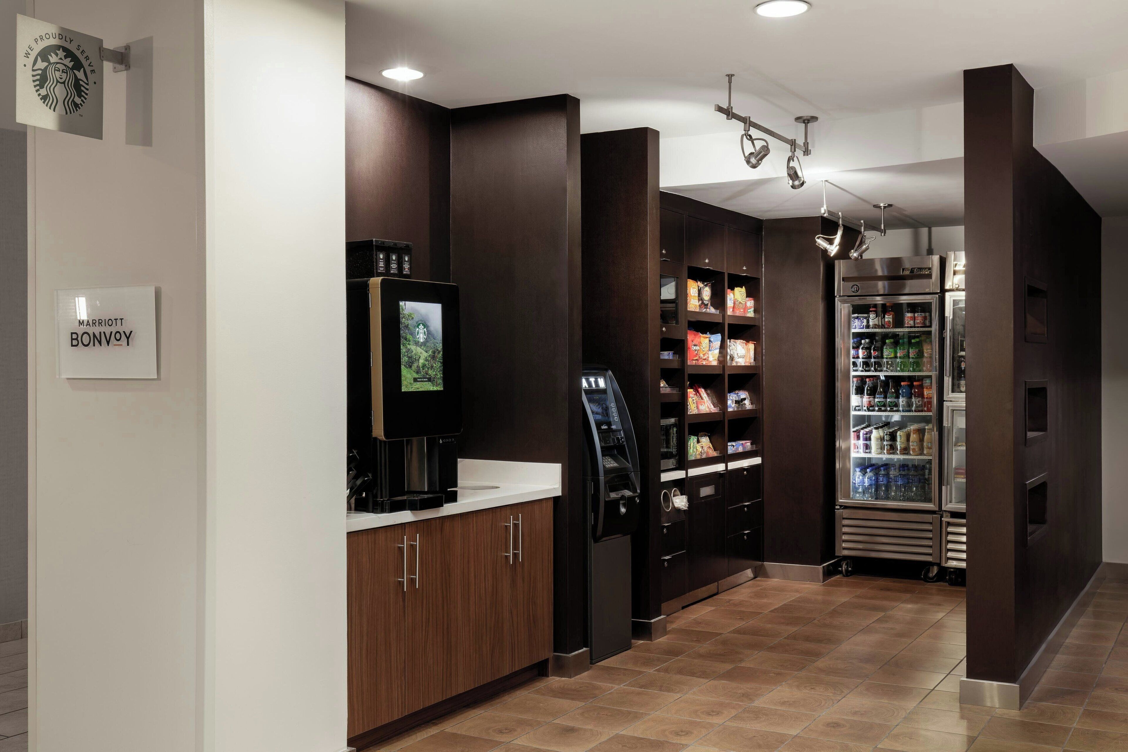 Un vestíbul d'hotel modern amb una màquina de cafè, una caixa d'autoservei i un dispensador de begudes refrigerades.