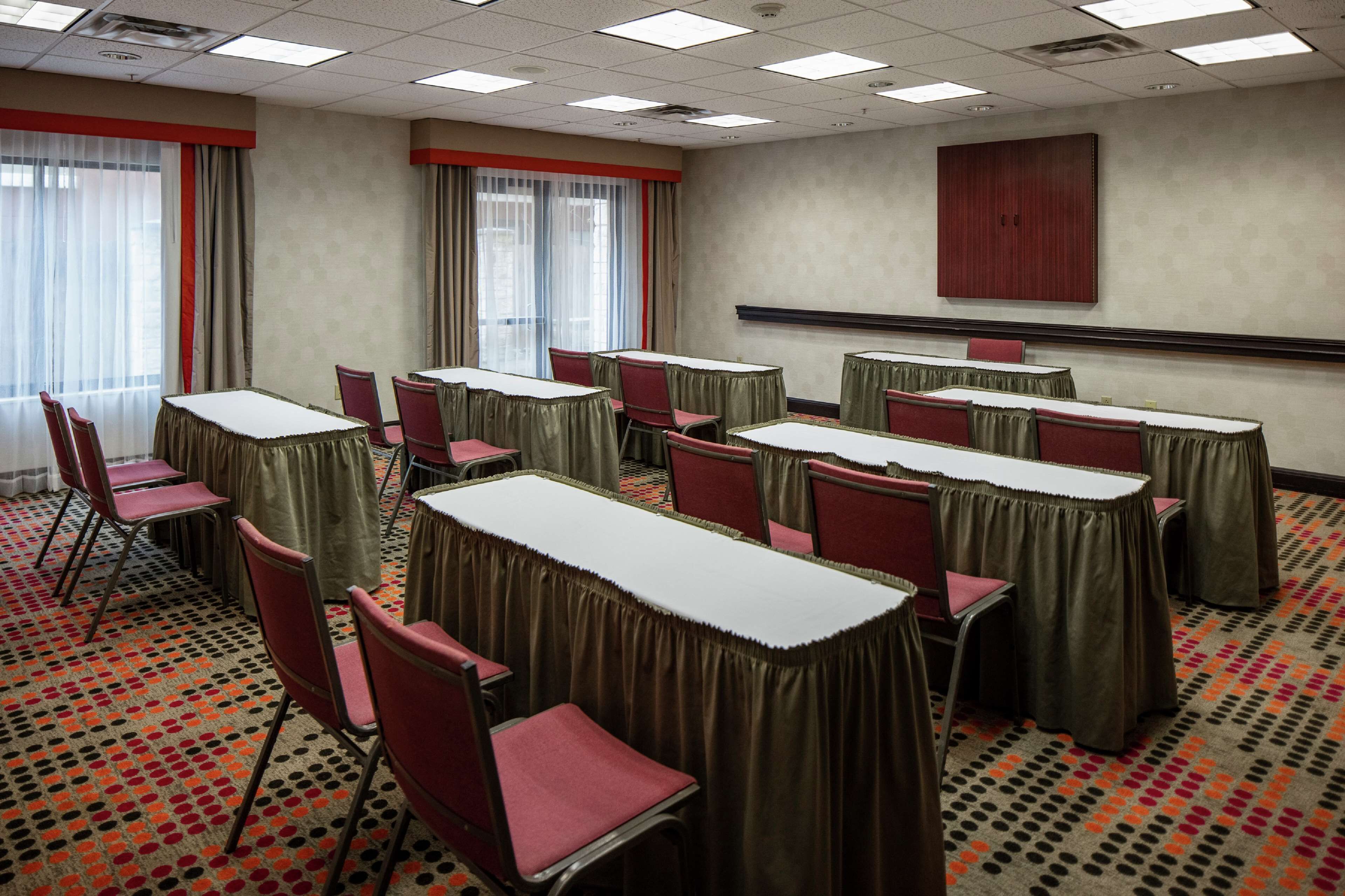 Une salle de conférence équipée de tables et de chaises, de grandes fenêtres avec des rideaux et d’un écran de projection mural.