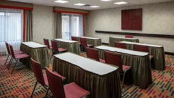 Une salle de conférence équipée de tables et de chaises, de grandes fenêtres avec des rideaux et d’un écran de projection mural.