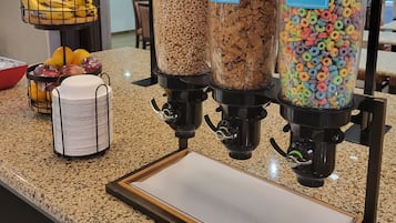 En frukostbuffé med tre behållare märkta med texten ”Kellogg’s Raisin Bran”, ”Kellogg’s Froot Loops” och ”General Mills Cheerios”. På disken står en skål med frukt och en vit behållare.