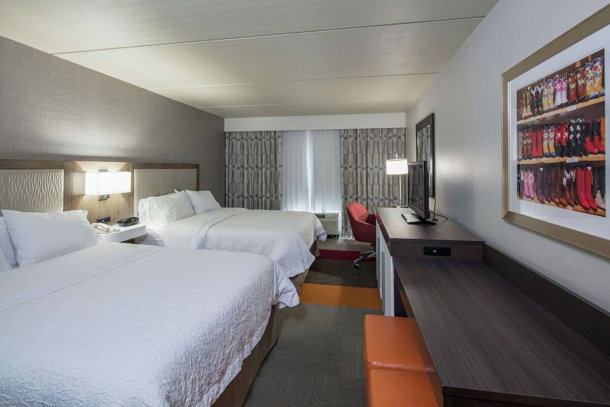 A hotel room with two beds, a desk, a chair, a TV, and a framed picture of boots.