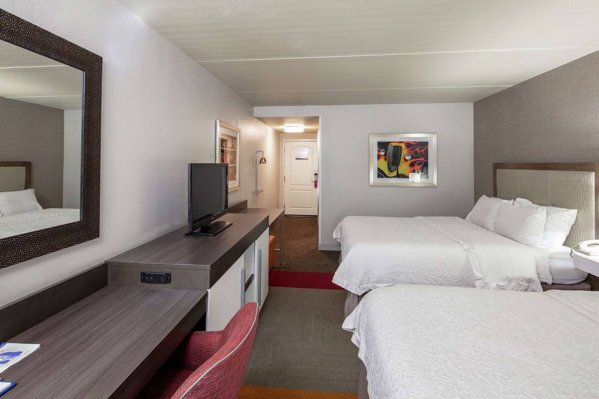 Habitación de hotel con escritorio, dos camas, televisor y espejo.