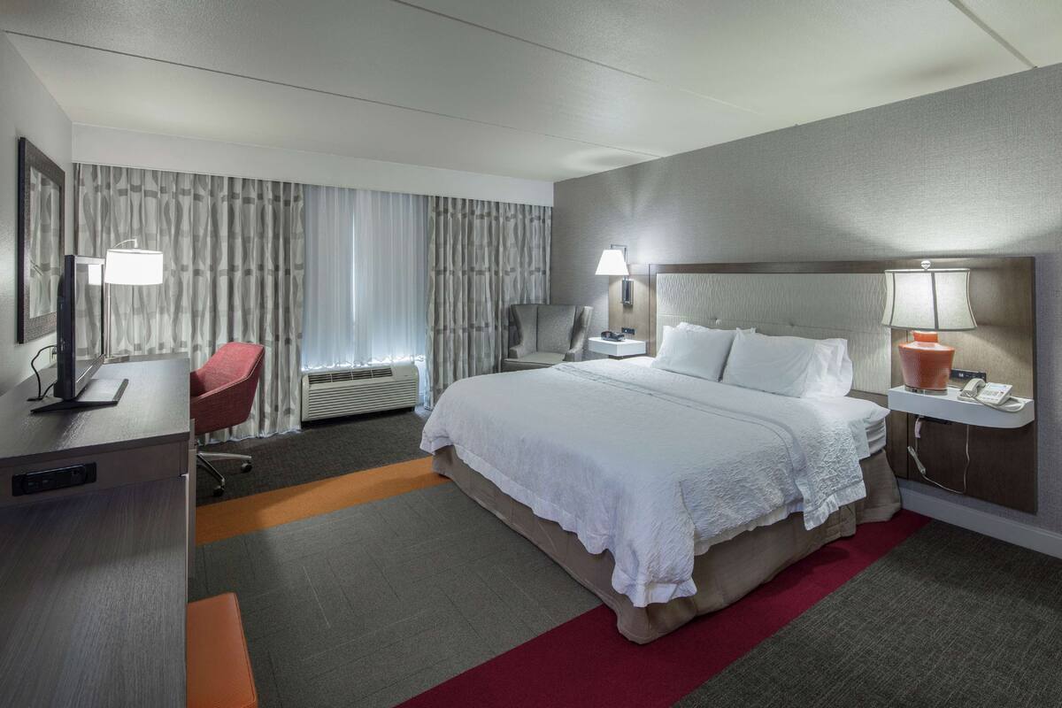 Una habitación de hotel con cama, escritorio, silla y un ventanal con cortinas.