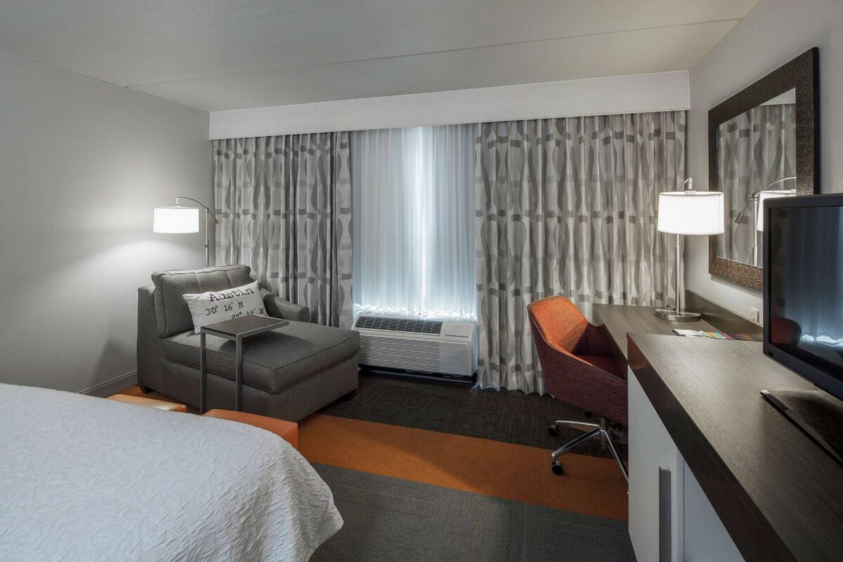 Habitación de hotel con cama, escritorio con televisor, silla, sofá y ventana con cortinas.