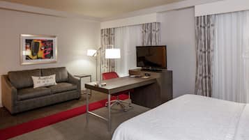 A hotel room with a bed, a desk with a TV, a sofa with a pillow, a lamp, and a framed picture on the wall.