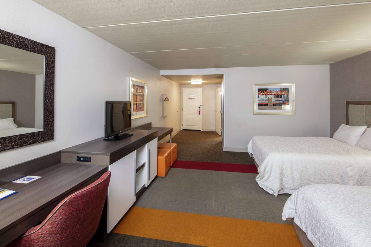 Habitación de hotel con escritorio, televisión, dos camas, espejo y alfombra roja.