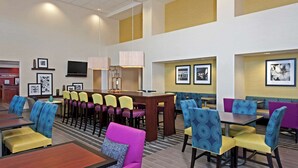 A hotel dining area with multiple tables and chairs, a central bar, and framed artwork on the walls. - Hampton Inn & Suites Bloomington - Normal (Normal)