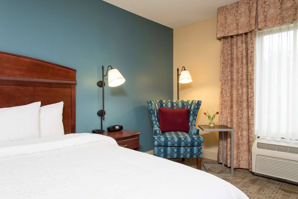 A hotel room with a large bed, a wooden headboard, two wall-mounted lamps, a patterned armchair, a small table with a vase, and a window with curtains.