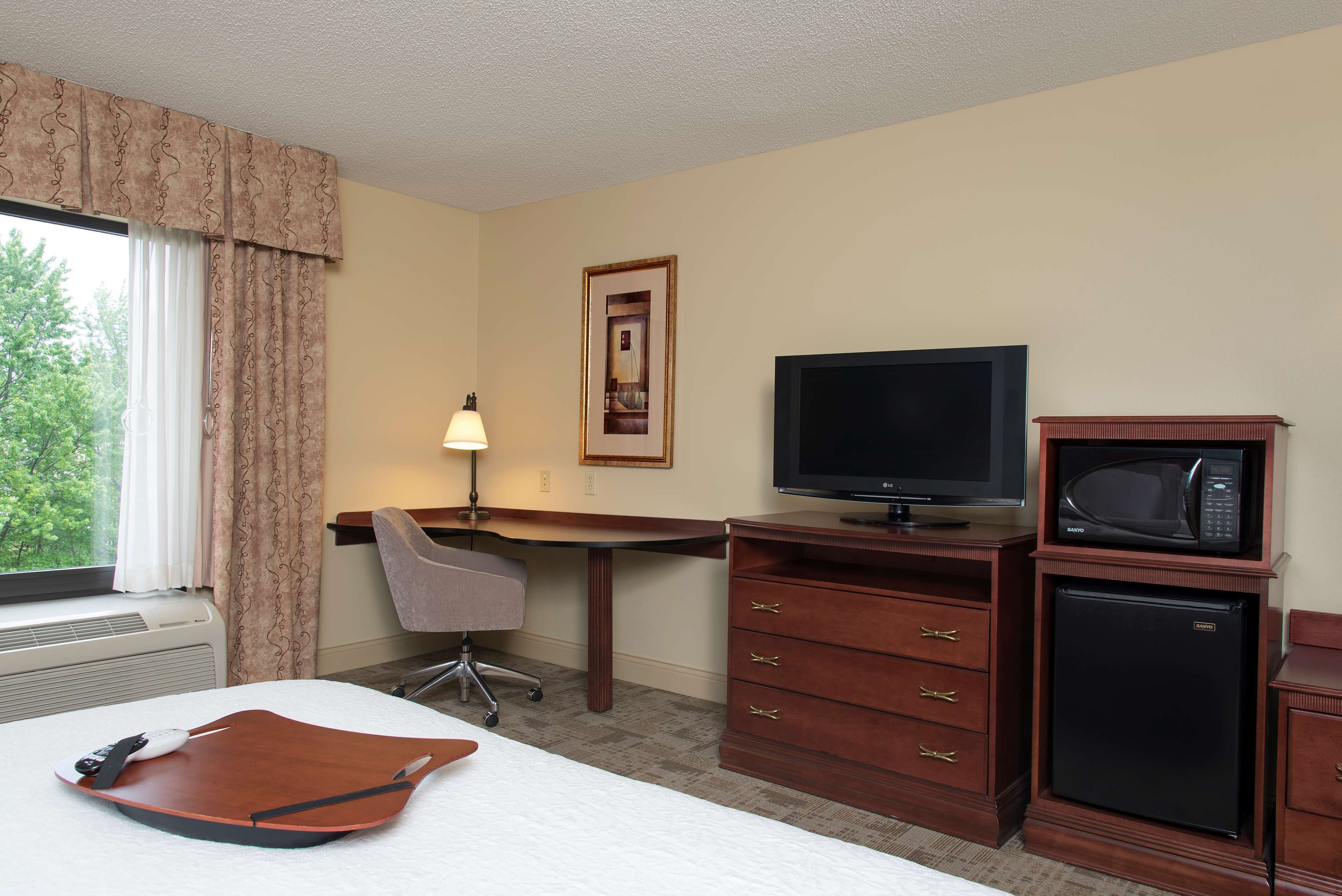 Ein Hotelzimmer mit Bett, Schreibtisch, Stuhl, Fernseher, Mikrowelle, Kühlschrank und einem fenster mit Vorhängen.