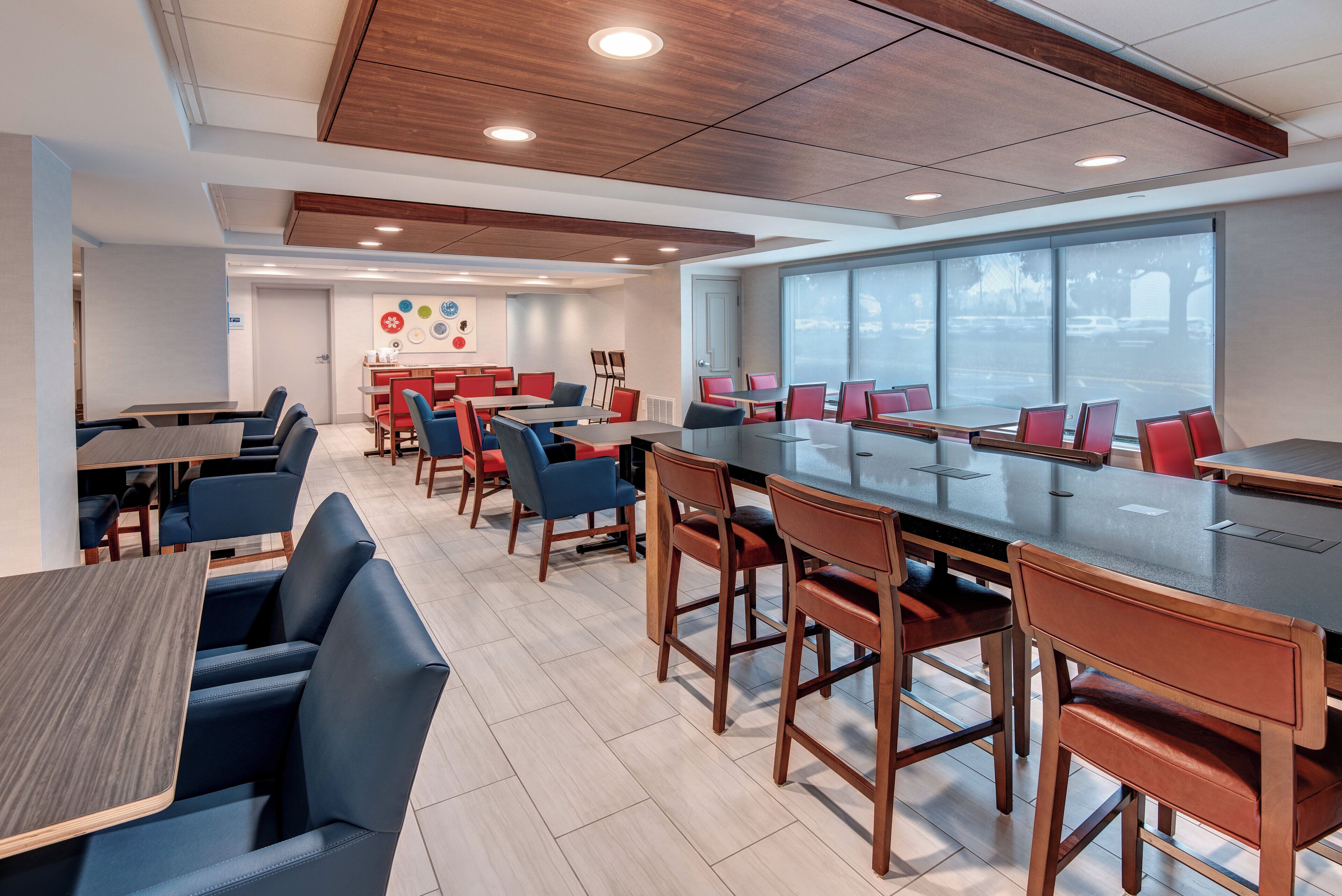 A modern dining area with multiple tables and chairs, a large window with a view, and a wall with decorative plates.