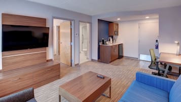 Une chambre d’hôtel moderne équipée d’un téléviseur à écran plat, d’un canapé, d’une table basse, d’un bureau avec une chaise, d’une kitchenette et d’un espace douche.