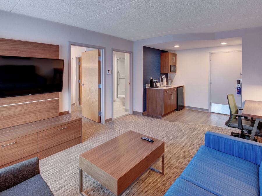 Une chambre d’hôtel moderne équipée d’un téléviseur à écran plat, d’un canapé, d’une table basse, d’un bureau avec une chaise, d’une kitchenette et d’un espace douche.