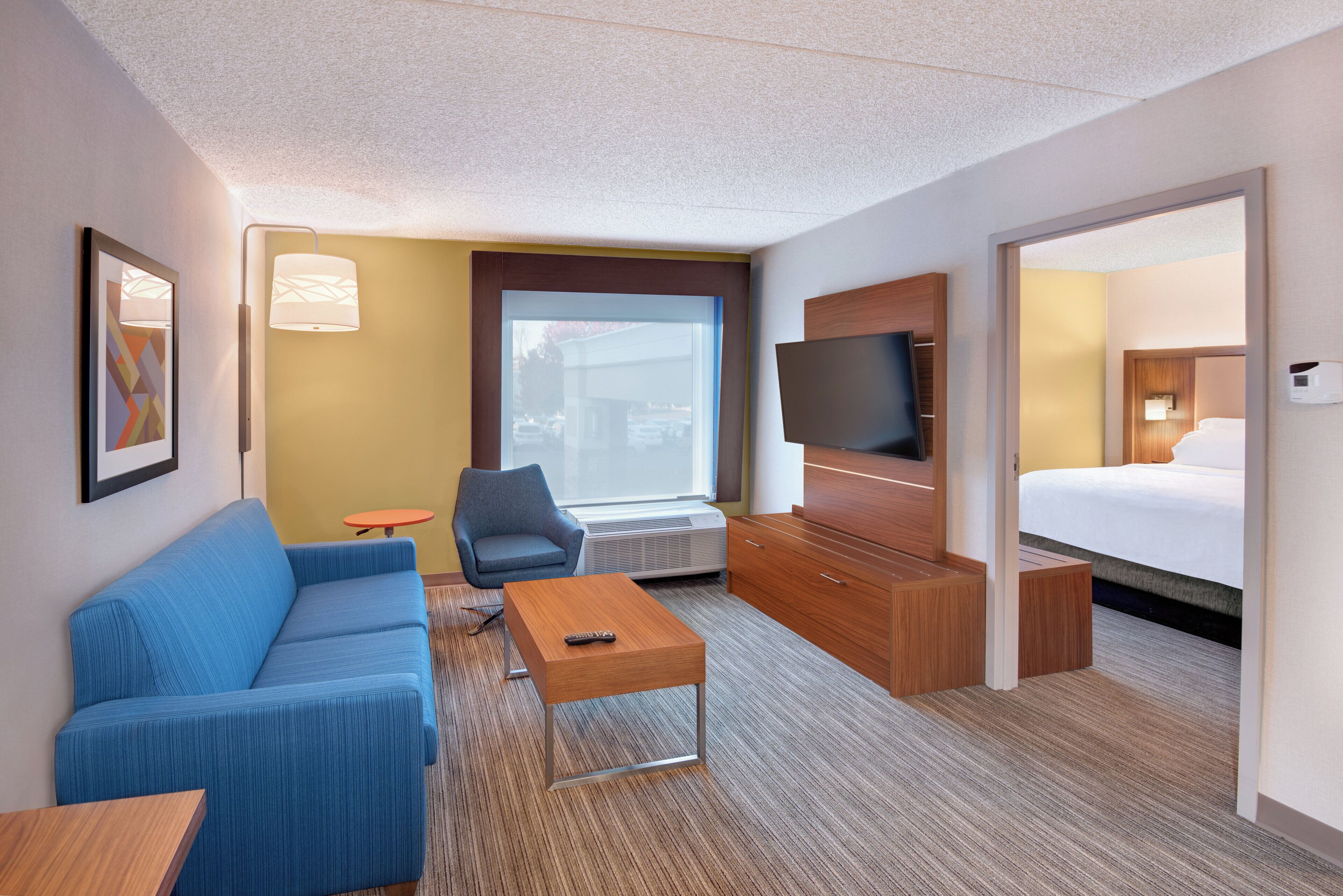 A hotel room with a blue sofa, a small table, a TV, a bed, and a window with a view of the outside.