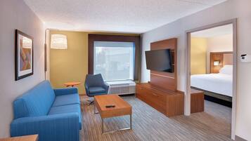 A hotel room with a blue sofa, a small table, a TV, a bed, and a window with a view of the outside.