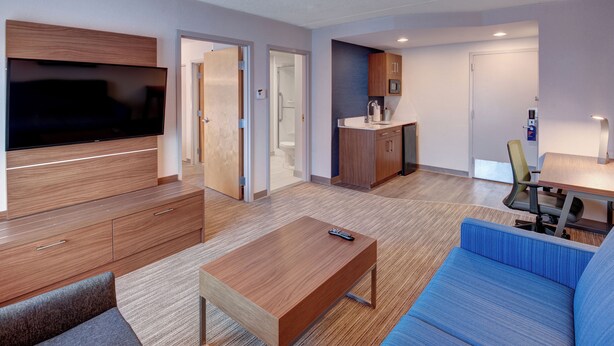 A modern hotel room with a flat-screen TV, wooden furniture, a blue sofa, a small table, and a kitchenette area.