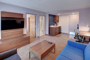 A modern hotel room with a flat-screen TV, wooden furniture, a blue sofa, a small table, and a kitchenette area.