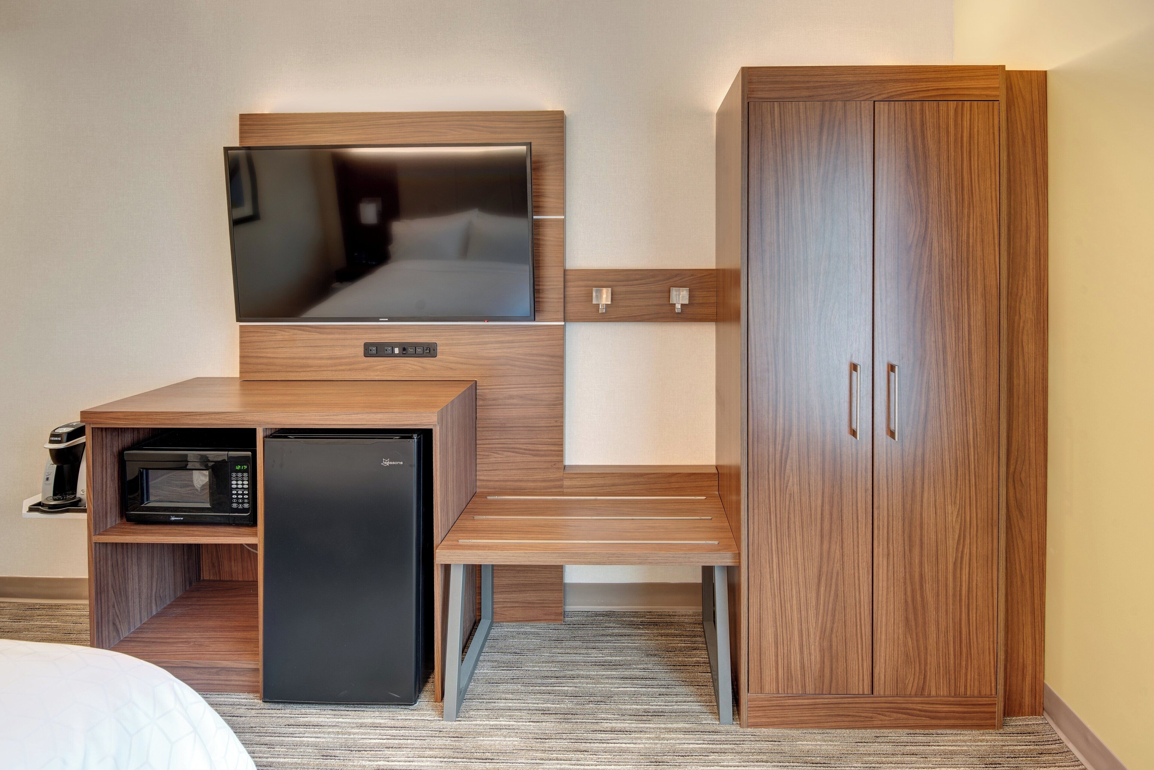 A compact hotel room with a flat-screen TV, a small refrigerator, a microwave, and a wooden wardrobe with two doors.