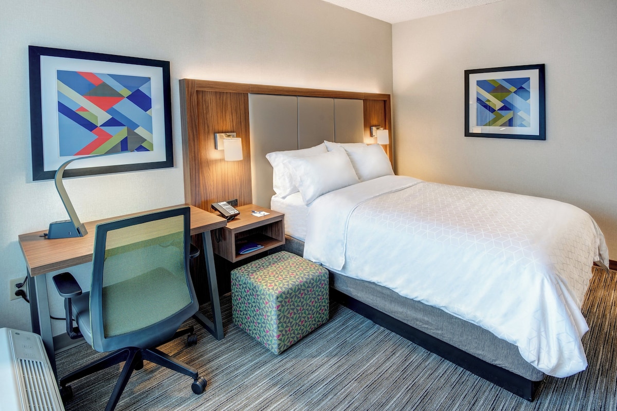 A hotel room with a bed, a desk with a chair, a nightstand, and two wall-mounted lamps.
