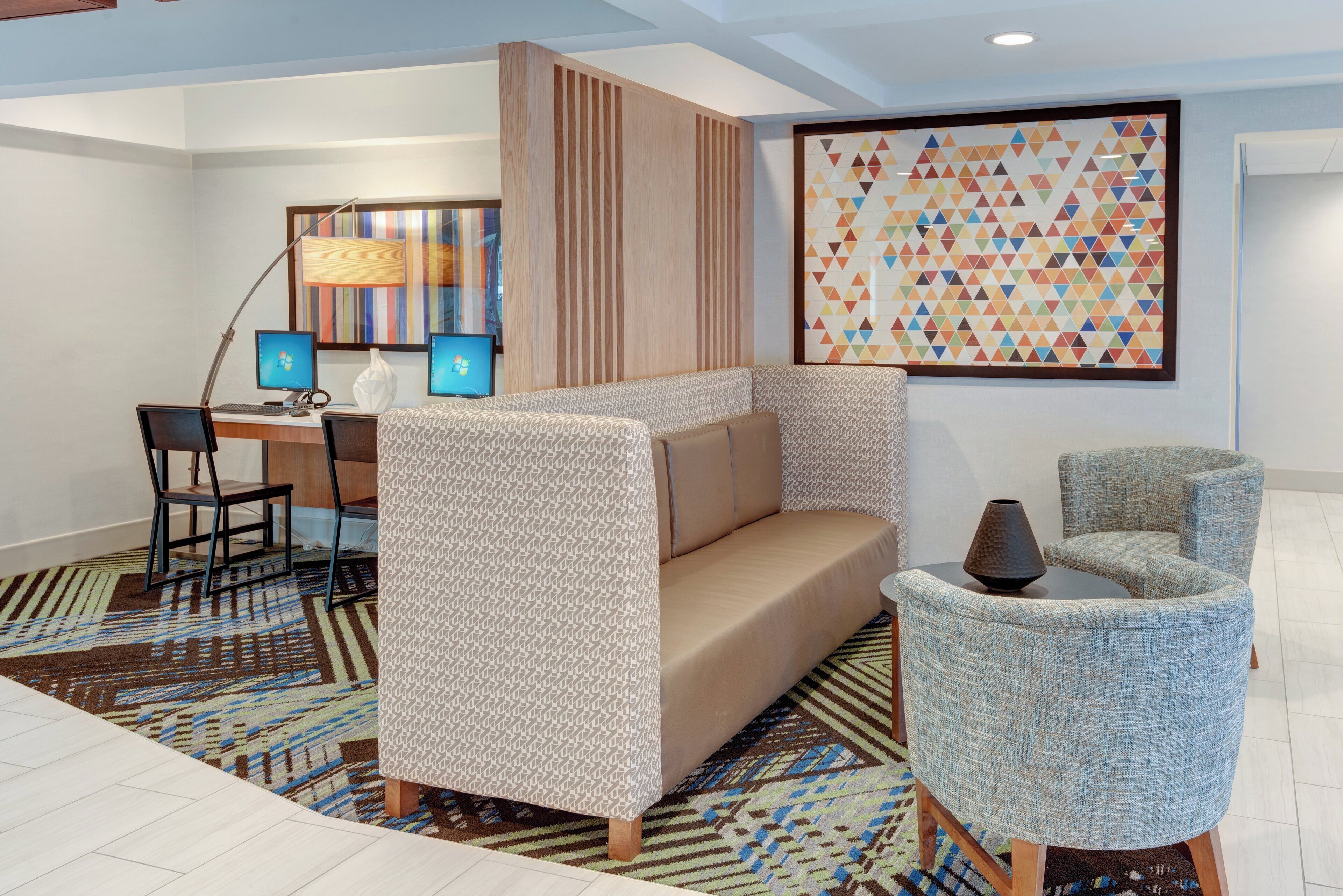 Una habitación de hotel moderna con sofá, dos sillones, un escritorio con computadoras y una gran obra de arte mural con diseño geométrico.