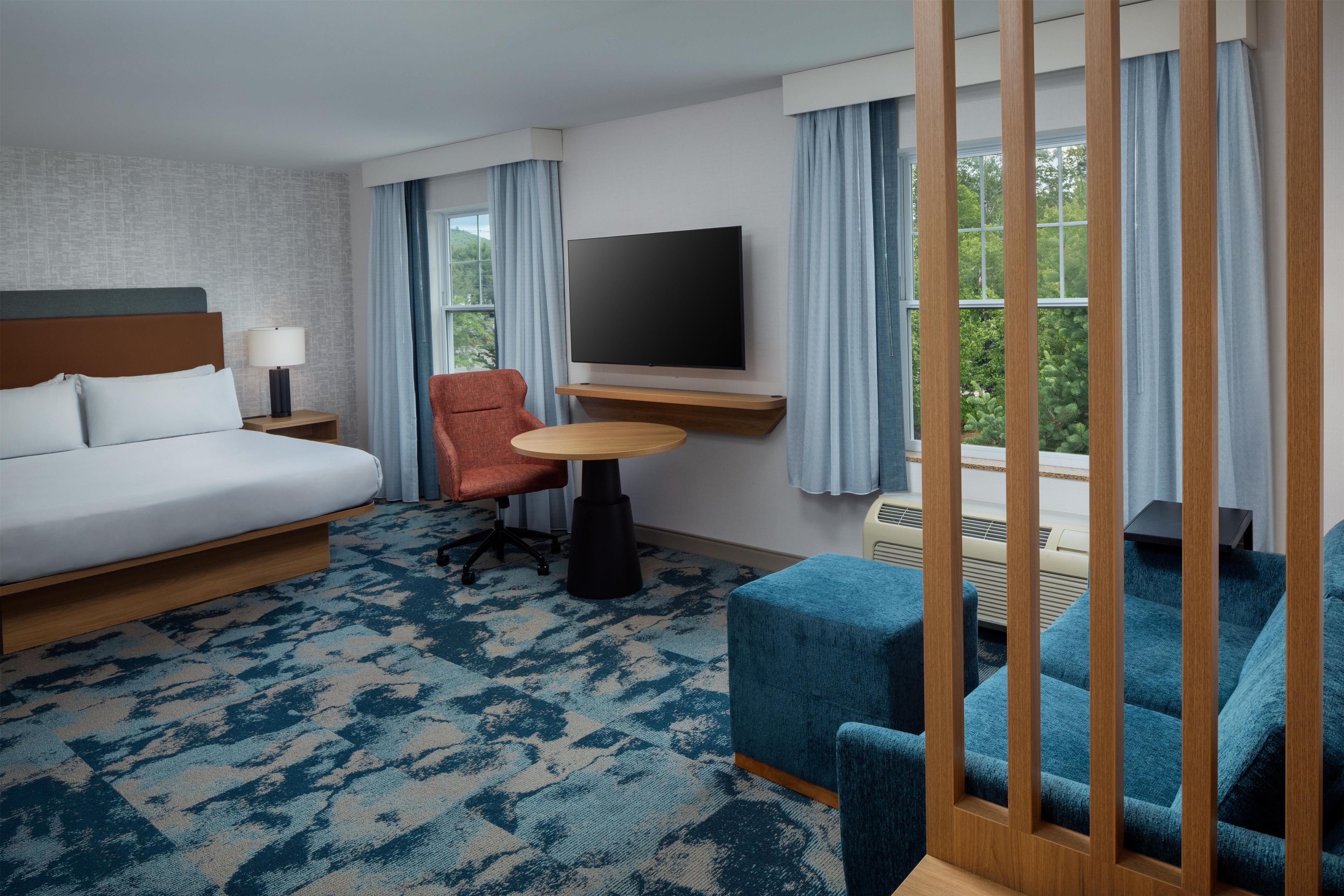A hotel room with a bed, a desk, a chair, a TV, and a view of greenery.