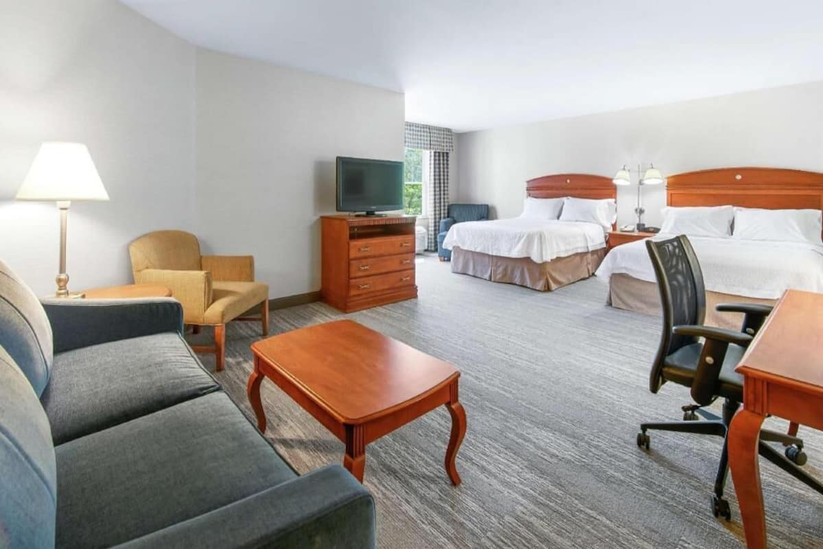A hotel room with a sofa, a wooden coffee table, a TV on a dresser, two beds with white linens, and a chair.