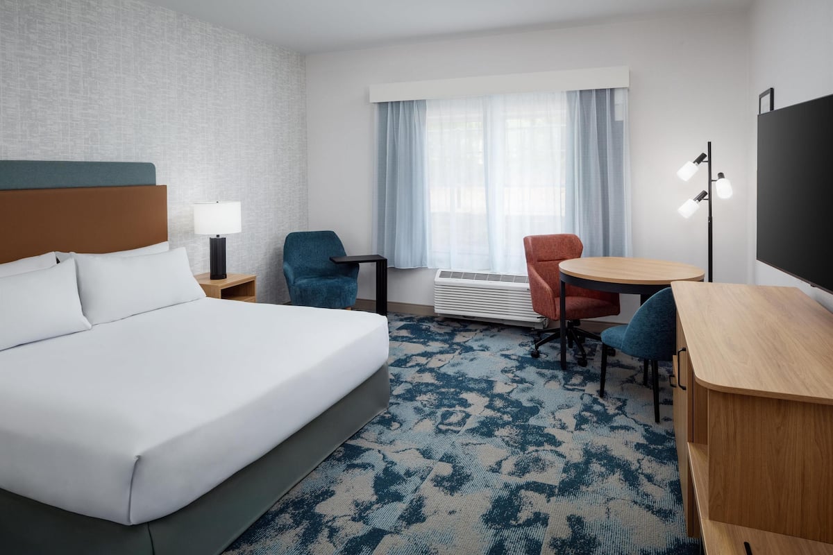 A hotel room with a large bed, a desk, two chairs, a lamp, a heater, and a window with curtains.